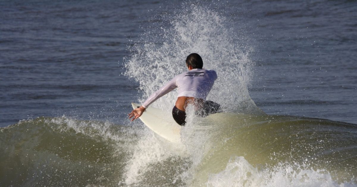 Imagem de um homem surfando, com uma prancha branca, para fazer referência ao fato de que a praia da Tereza possui ondas ideias para quem surfa