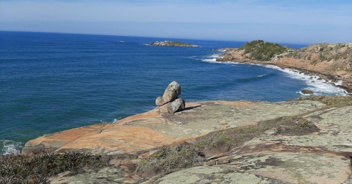 Imagem de um mar, com pedras ao redor, para fazer referência trilha que leva até a praia da Ilhota, que possui cerca de 3,7 km ida e volta