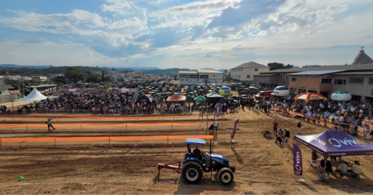 Imagem de uma estrada de terra com tratores, ao redor de barraca e pessoas, para mostrar um dos pontos principais da festa religiosa, o desfile de tratores de Treze de Maio 