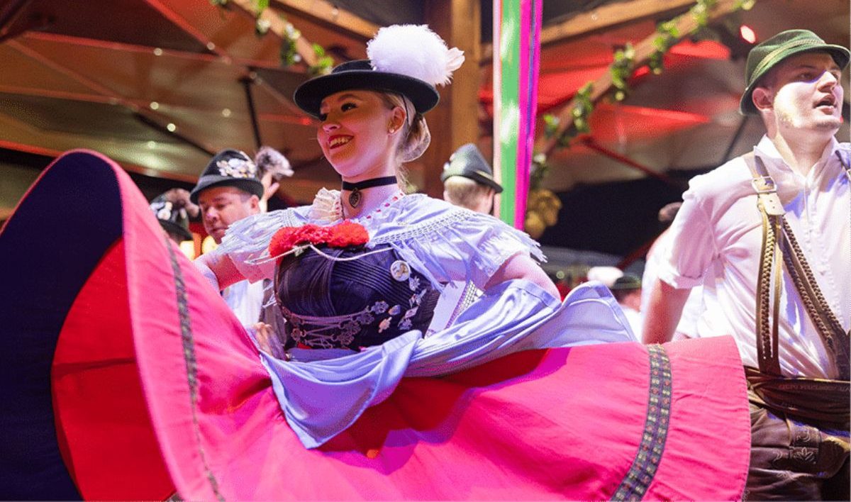 Foto horizontal mostra uma mulher jovem, de pele clara, rosto arredondado e cabelos loiros presos. Ela sorri, está com as duas mãos na cintura e está vestida com um traje típico. Sua saia é vermelha e está rodando. Ela também usa um chapéu preto com uma pluma branca. Do lado direito, um homem jovem de pele clara também veste um traje típico. Ele usa um chapéu verde, uma blusa de manga curta branca, uma bermuda marrom e um suspensório verde. Eles estão dentro de um salão. Ao fundo aparecem algumas pessoas.