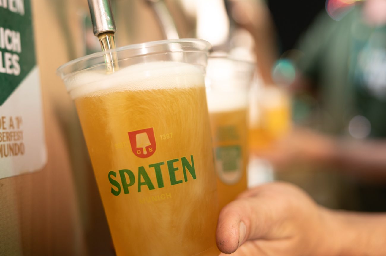 Foto horizontal mostra parte de uma mão de pele clara, segurando um copo de plástico cheio de cerveja com a logomarca da Spaten. Ele está debaixo de uma torneira de chopp aberta. O fundo é desfocado.