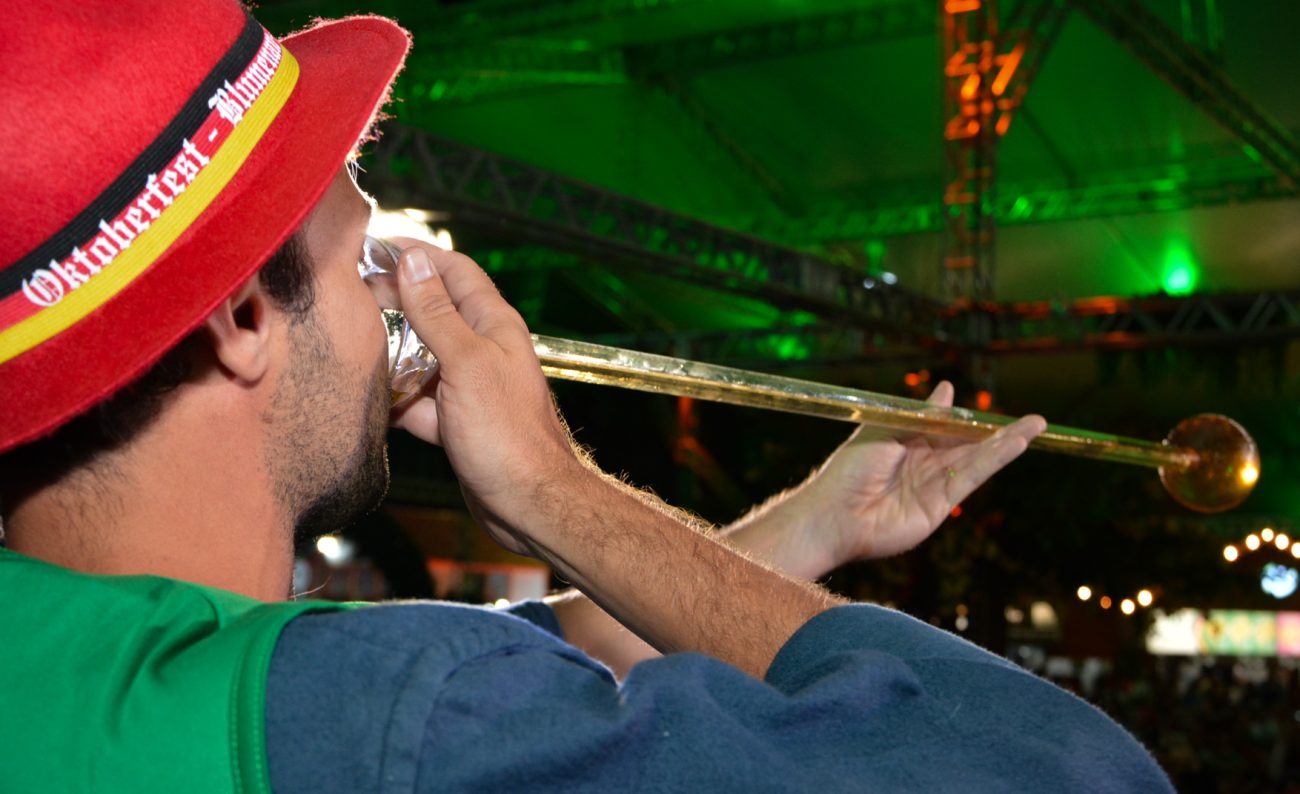 Foto horizontal de um homem jovem, de pele clara, barba preta cerrada e cabelo curto preto. Ele usa um chapéu vermelho escrito Oktoberfest Blumenau e segura próximo à boca uma longa taça de vidro com chope dentro.