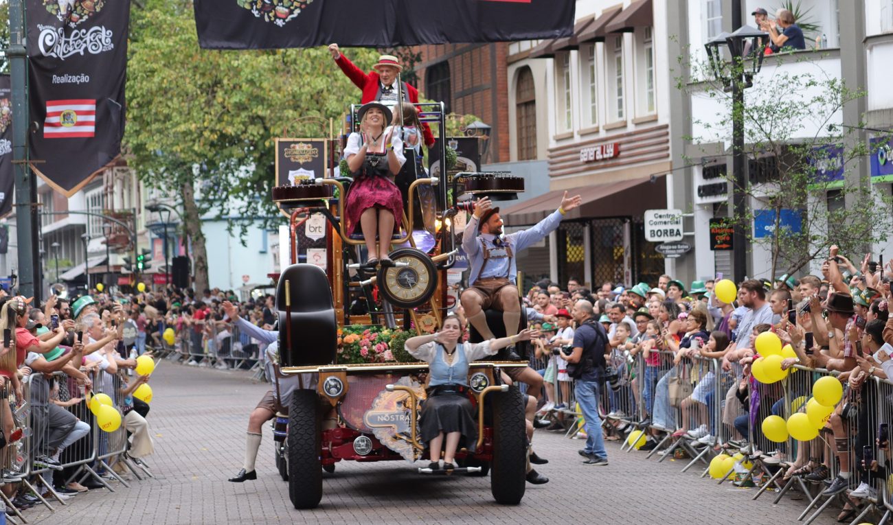 Foto mostra um carro alegórico com cinco pessoas usam trajes típicos passando pela Rua XV de Novembro, durante a Oktoberfest em Blumenau. Nas laterais da via há várias grades metálicas, onde se apoiam milhares de pessoas que acompanham o desfile. Ao fundo, prédios históricos da rua e uma árvore.