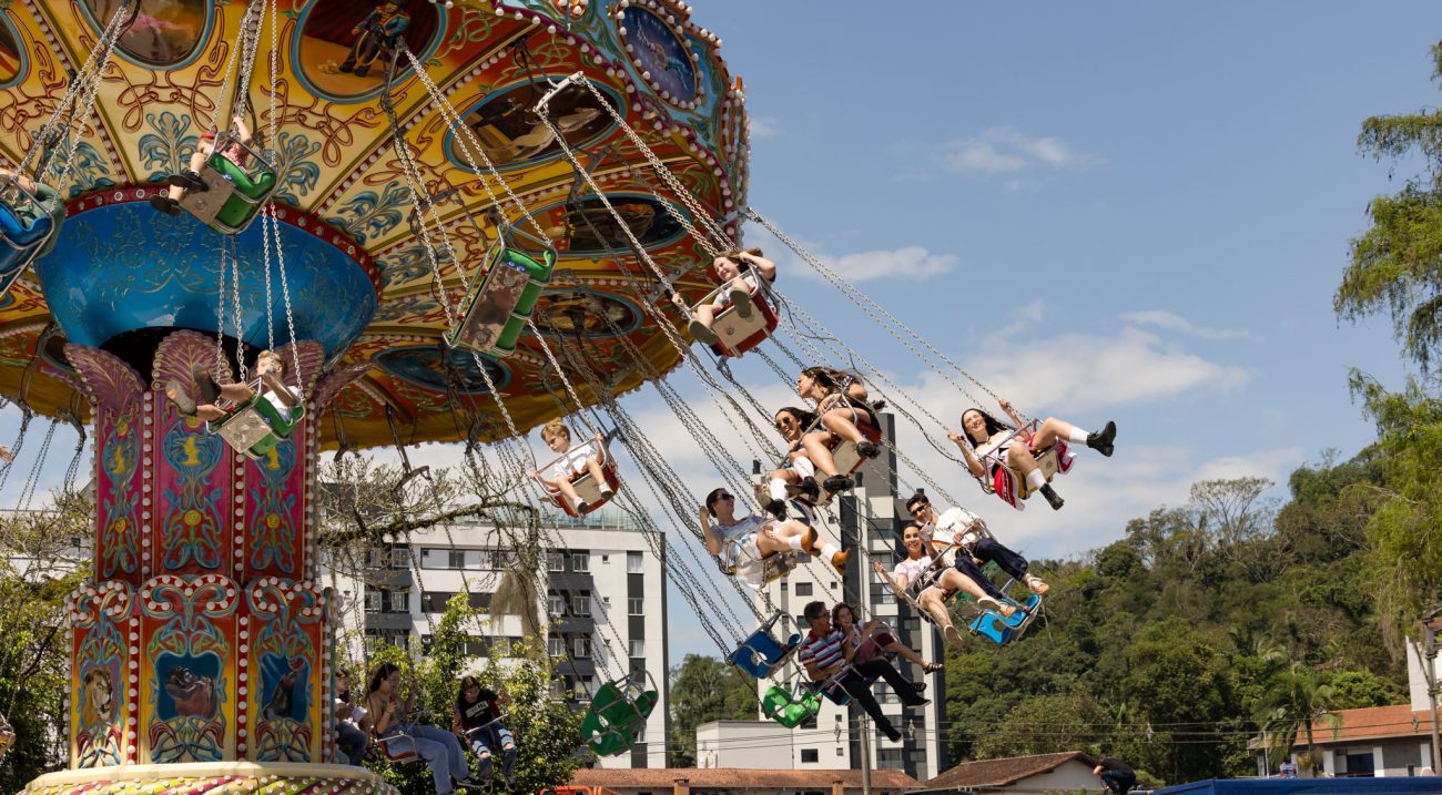 Foto horizontal mostra 15 pessoas sentadas em balanços aéreos de um carrossel todo colorido e ricamente decorado. Ao fundo, aparecem dois prédios e algumas árvores. O céu está bem azul.