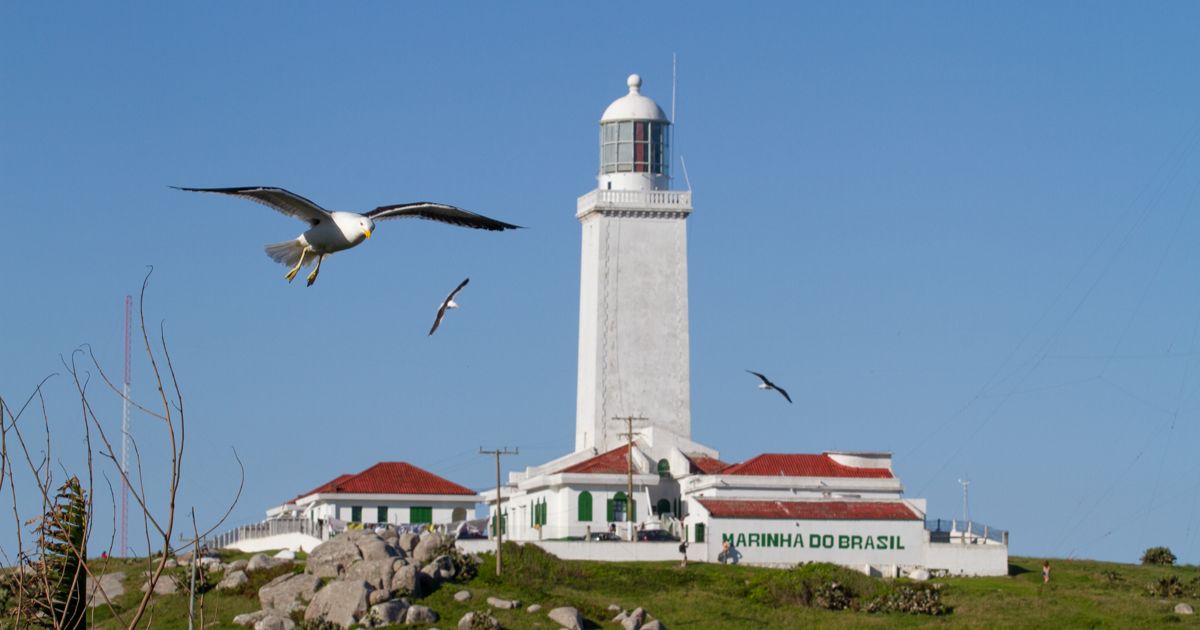 Imagem do farol de Santa Marta visto de longe, com o pátio dele em destaque, com uma gaivota vando, para fazer referência ao fato de que a Parte externa do maior farol das Américas está sempre aberta para visitação