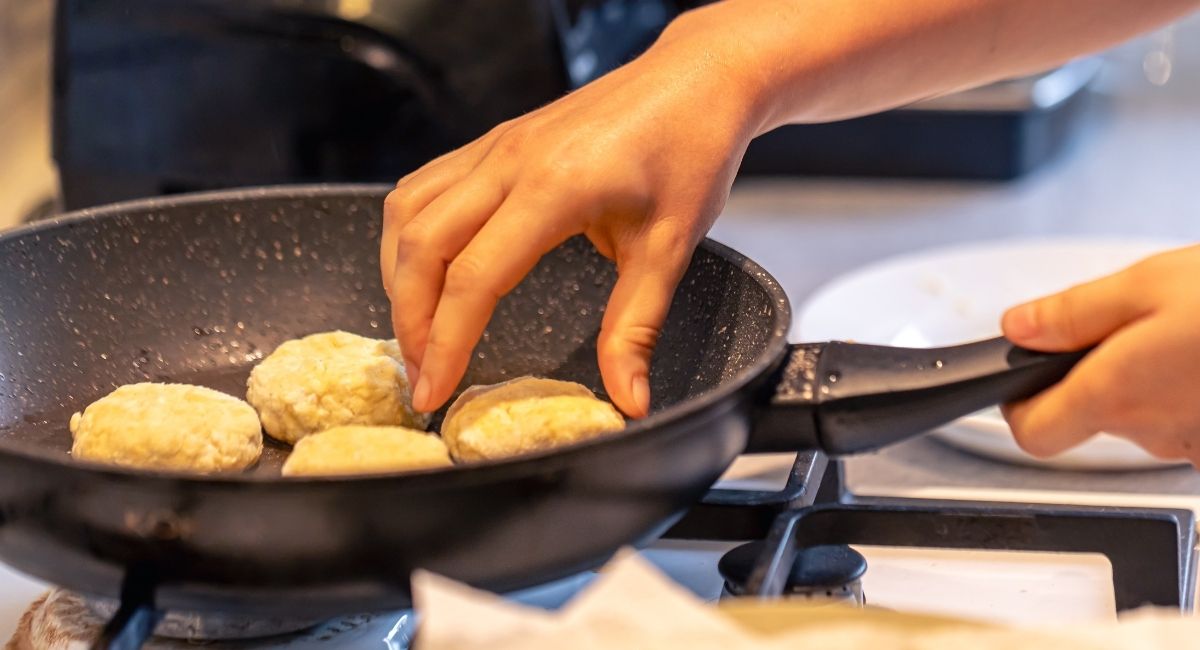 Mãos de uma pessoa fritando bolinhos em uma frigideira sobre o fogão, com óleo quente, cena que representa o preparo de alimentos e o cheiro de fritura espalhando pela cozinha