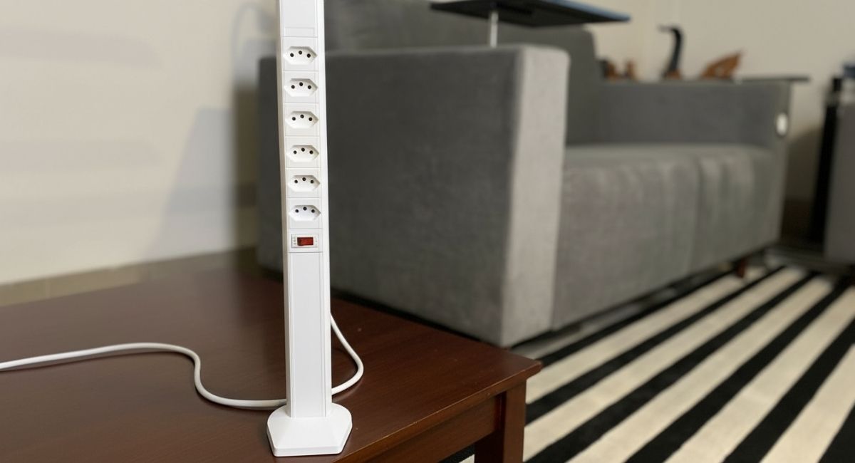 Filtro de linha branco extensível em formato vertical, com várias tomadas alinhadas e botão liga/desliga vermelho na parte inferior, apoiado sobre uma mesa de madeira. Ao fundo, há um sofá cinza e um tapete listrado preto e branco em uma sala de estar