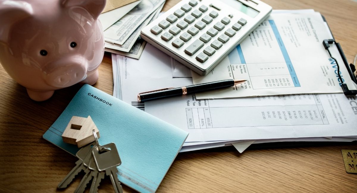Mesa com documentos financeiros, calculadora, caneta, um porquinho de cofre, chaves e um caderno, representando controle de gastos, impostos e organização financeira conforme a lei do aluguel.