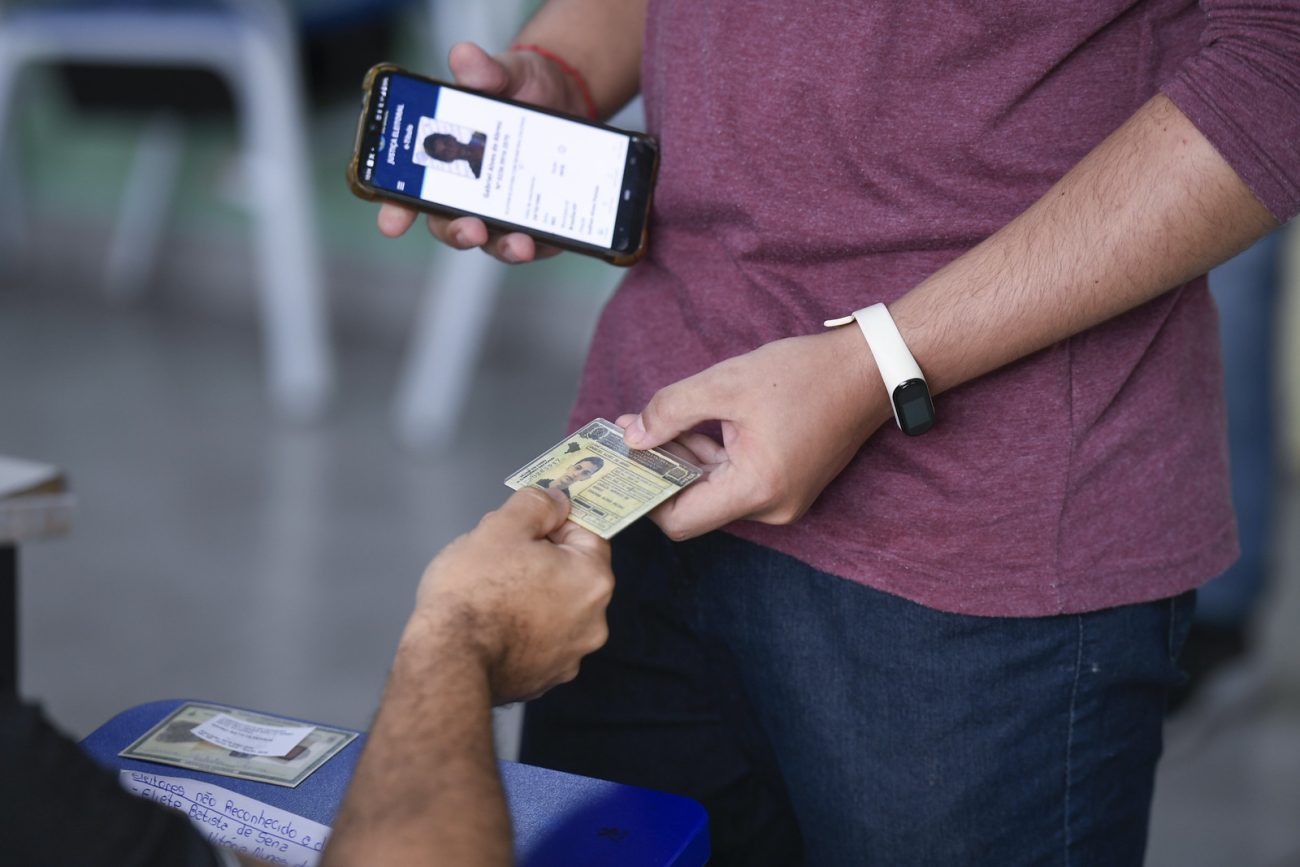 homem mostra carteira de motorista e segura celular com aplicativo e-título aberto para mesário. ilustra matéria sobre eleições 2026.