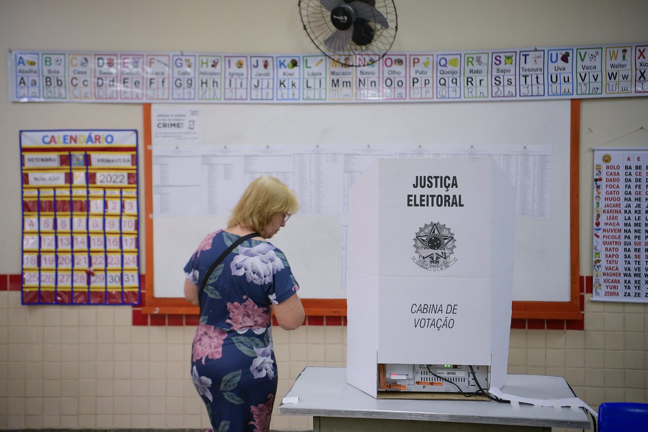 uma mulher de cabelos louros e vestido florido se direciona a uma cabina de votação em uma sala de aula. ilustra matéria sobre eleições 2026.