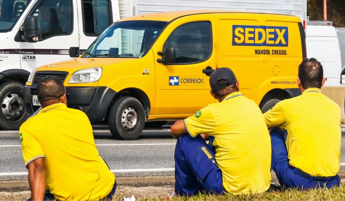 Três trabalhadores dos Correios sentados diante de um veículo do Sedex; imagem ilustra notícia sobre a greve dos Correios