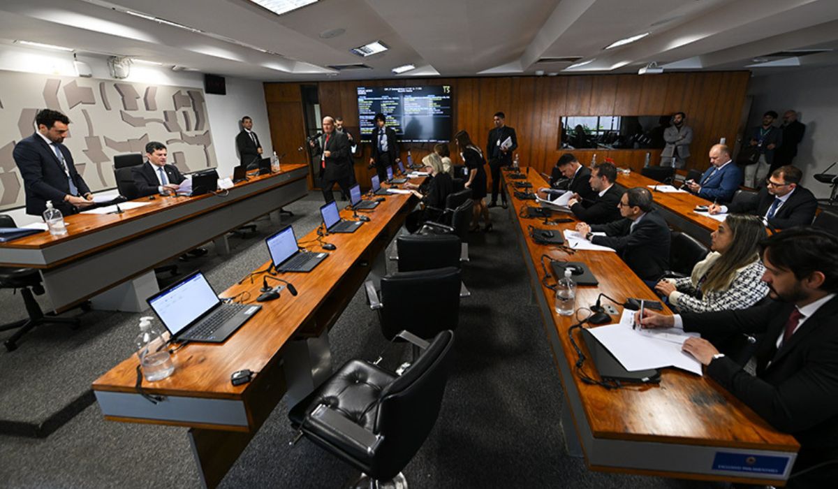 Parlamentares e outros participantes reunidos em sala de comissão da Câmara dos Deputados, com computadores, microfones e documentos sobre as mesas