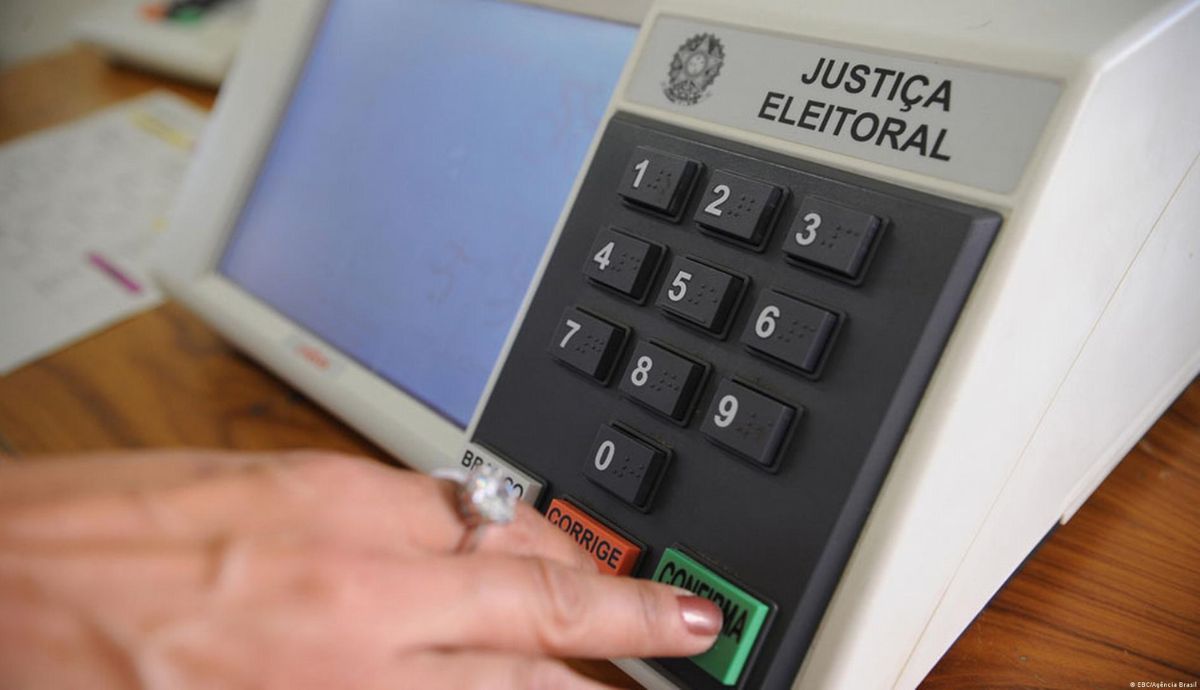 Eleitor aperta o botão verde de confirmação em uma urna eletrônica, com teclado numérico e identificação da Justiça Eleitora