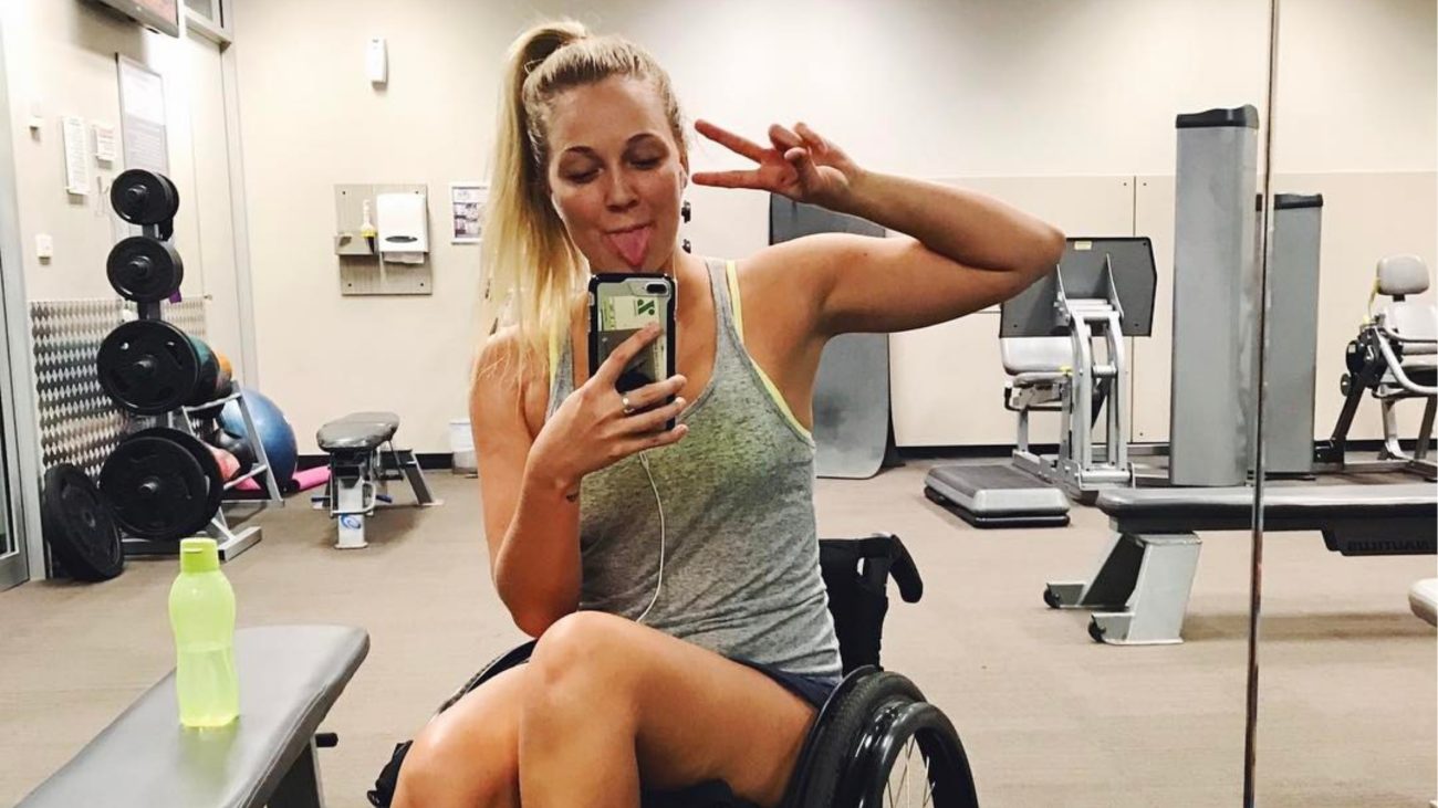 Paraquedista aparece em uma academia durante um momento de recuperação, sentada em uma cadeira de rodas e fazendo uma pose para a foto.