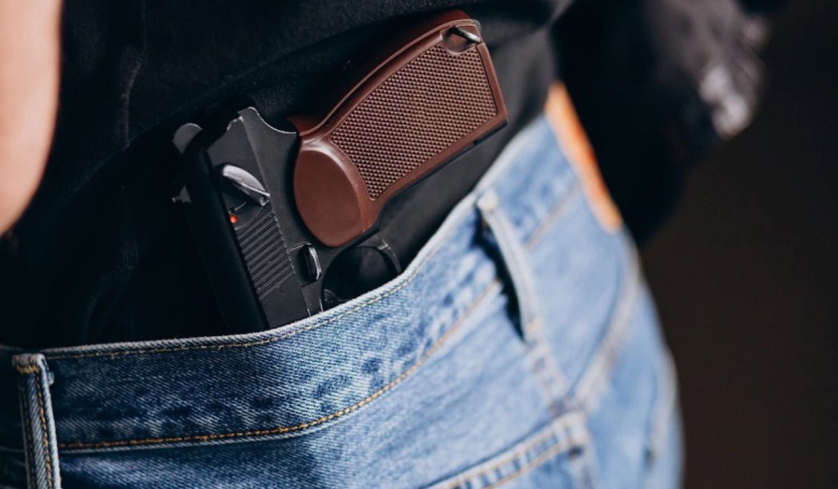 Arma de fogo com cabo marrom posicionada na cintura de uma pessoa que veste calça jeans e camiseta preta
