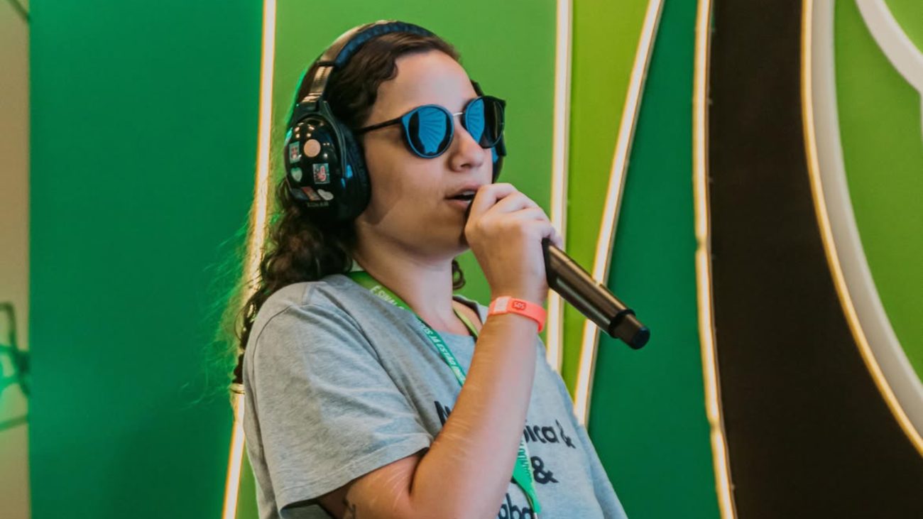 Fernanda Fialho aparece durante um evento, usando fones de ouvido e óculos escuros enquanto segura um microfone.
